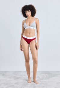 Hallutsuhhallise halli puuvillane bralette õhukeste rihmadega, millel on valge vöökoht. Punane ribakudumiga string, millel on valge logo detailid. Modell seisab paljajalu helesinisel vaibal.