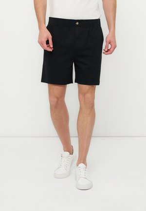 Homme portant un short à taille élastique noire et des baskets blanches, debout devant un fond blanc uni.