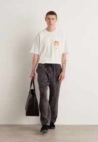 Hvit bomull T-skjorte med oransje fruktdesign, kombinert med grå teksturerte bukser, svart totebag og mørke sko mot en nøytral bakgrunn.