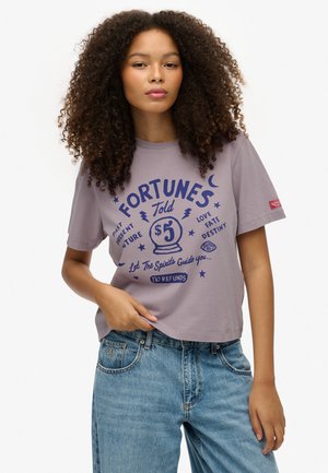 Lilla bomuld t-shirt med et crop-top design. Har fed blå tekst og grafik relateret til spådomskunst. Slidte blå denimjeans.
