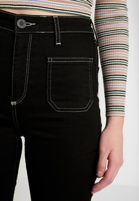Svarta jeans med hög midja i denim med kontrasterande vit söm, en klassisk knappstängning och en enda bakficka med rektangulär form.