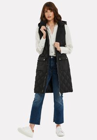 Schwarze gesteppte Weste mit Kapuze, Reißverschluss und Druckknöpfen. Mit seitlichen Taschen und einer langen, lockeren Passform. Getragen über einem weißen Hemd und Jeans.