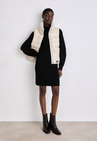 Colete felpudo bege com gola alta e fecho de zip, vestido sobre um vestido preto de malha canelada, combinado com botas de tornozelo castanhas escuras. Texturas suaves.