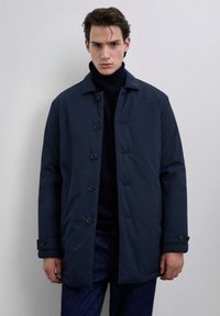 Hombre vistiendo un abrigo oscuro azul marino abotonado sobre un suéter negro de cuello alto, de pie contra un fondo gris claro uniforme.