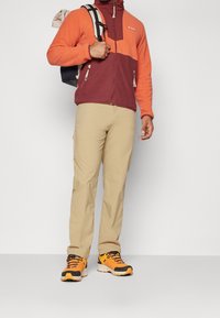 Giacca in pile arancione e bordeaux con zip, pantaloni beige e scarpe da ginnastica arancioni/grigie. Include uno zaino con un design a scacchi.