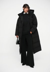 Zwarte pufferjas met een lange silhouette en een boxy ontwerp, gecombineerd met een zwarte trui, brede broek en een zwarte handtas.
