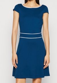 Robe ajustée bleue à manches courtes, avec un encolure dégagée et une bande d'accent blanche à la taille. Tissu lisse avec un bas évasé.