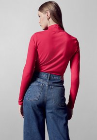 Rood long-sleeve top met hoge hals, gedragen met hoogzwart blauwe denim jeans met grote achterzakken en een relaxed fit.