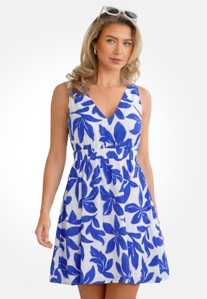 Kobieta ubrana w białą sukienkę bez rękawów z niebieskimi wzorami liści, złote kolczyki koła, wielowarstwowe naszyjniki oraz białe okulary przeciwsłoneczne na głowie.