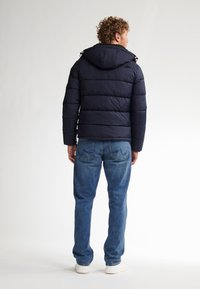 Chaqueta acolchada navy con capucha, diseño acolchado y puños elásticos, combinada con jeans azul claros y zapatillas blancas. Vista trasera.