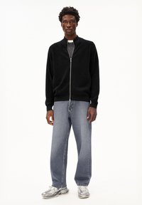 Jonge man die staat, gekleed in een zwarte zip-up sweater, vervaagde blauwe jeans en wit-grijze sneakers, tegen een effen witte achtergrond.