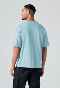 Άνδρας με κοντά σκούρα μαλλιά, που φοράει ανοιχτό μπλε κοντομάνικο φανελάκι και μαύρο παντελόνι cargo, στέκεται με την πλάτη γυρισμένη προς το απλό φόντο.