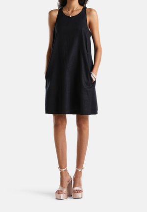 Femme portant une robe noire sans manches longueur genoux, coupe évasée, avec poches et des talons compensés couleur nude, debout devant un fond uni.