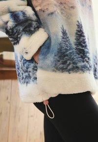 Fleece pullover with blue and white tree pattern, featuring a pocket and pink drawstring accent. Soft texture, relaxed fit, knee-length.