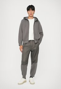 Felpa grigia con zip e cappuccio, T-shirt bianca sotto, pantaloni grigi e grigio scuro, sneakers bianche. Design minimalista, vestibilità comoda.