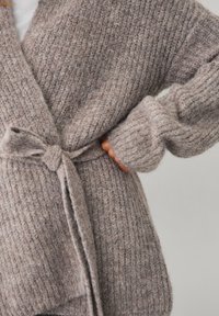 Frau in einer grob gestrickten taupefarbenen Strickjacke, die an der Taille gebunden ist, mit einer Hand, die unter dem Ärmel an ihrer Hüfte steckt.