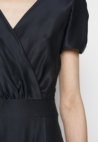 Detailní záběr osoby oblečené v černých saténových šatech s krátkým rukávem, zavinovacím střihem, výstřihem do V a elastickým pasem.