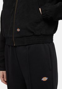 Person wearing black Dickies corduroy jacket with zipped front and matching black pants with Dickies logo on the pocket.