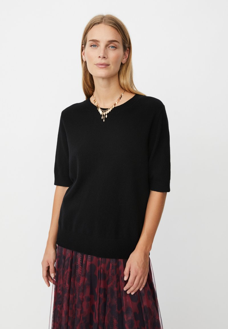 Ung kvinde med blont hår iført en sort kortærmet sweater, et guldbladformet halskæde og en rød mønstret nederdel.