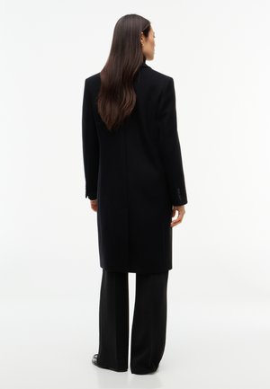 Femme aux longs cheveux foncés porte un manteau noir jusqu’aux genoux et un pantalon large noir, debout de dos.