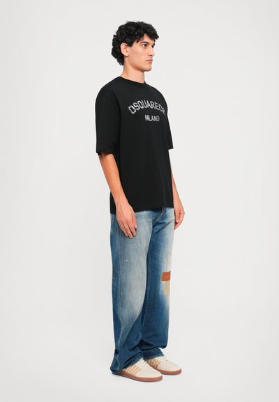 Giovane uomo in una maglietta nera Dsquared2 Milano, jeans blu sbiaditi con toppe e sneaker bianche, di fronte a uno sfondo bianco semplice.