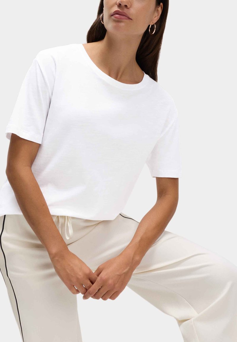 Weißes T-Shirt mit kurzen Ärmeln aus weichem Stoff, mit Rundhalsausschnitt und lockerem Schnitt. Kombiniert mit hellfarbenen, mit Kordelzug versehenen Hosen.