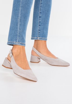 Femme portant des chaussures à talons blocs à bride arrière en daim gris clair, à bouts pointus, associées à un jean skinny bleu sur un sol blanc.