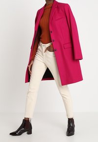 Manteau en laine magenta au design ajusté, avec deux grandes poches, associé à un haut marron côtelé et un jean blanc à ourlet effiloché, le tout complété par des bottines noires.