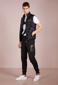 Crna podstavljena prsluk s patentnim zatvaračem, bijela majica s grafikom, crne trenirka hlače s logom i bijele tenisice s crvenim detaljima. Glatka tekstura.