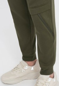 Olijfgroene sweatpants met ritsdetails aan de zijkant, tapered fit en een zachte textuur, gecombineerd met beige sneakers met gaasaccenten.
