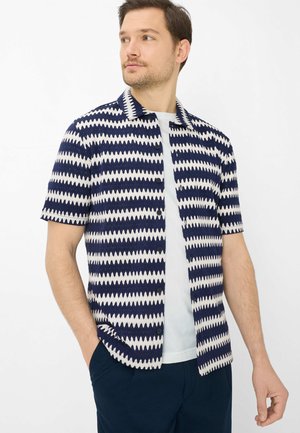 Mann trägt kurzärmeliges Hemd mit marineblauem und weißem Zickzackmuster über einem weißen T-Shirt, Hände in den Taschen einer marineblauen Hose, schaut nach links.