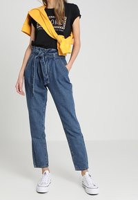Pantalones de denim de cintura alta con un ajuste holgado, color azul claro, cinturón de lazo y puños enrollados, combinados con una camiseta gráfica negra y zapatillas blancas.