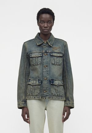 Kobieta w wyblakłej niebieskiej kurtce dżinsowej z czterema przednimi kieszeniami z klapami oraz jasnych spodniach na jednolitym białym tle.