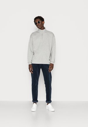 Sweatshirt cinza claro com fecho éclair e gola alta, combinado com calças de veludo cotelê azul-marinho e ténis brancos. Acessórios incluem óculos de sol escuros.