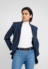 Blazer a righe navy con linee blu, vestibilità tailoring, spalline, indossato sopra un dolcevita bianco a costine e jeans in denim blu chiaro.