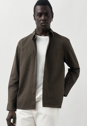 Mann trägt ein offenes braunes Hemd über einem weißen T-Shirt und weißen Hosen und steht vor einem schlichten hellen Hintergrund.