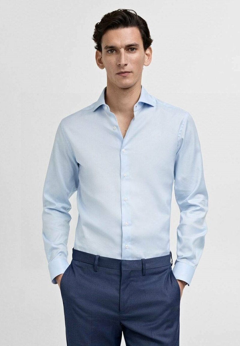 Cămașă albastră deschis cu mânecă lungă, guler ascuțit și închidere cu butoni, fabricată dintr-un material neted, asortată cu pantaloni negri.