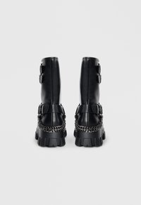 Botas de tobillo de cuero negro con suela gruesa, detalles de doble hebilla en el eje y una cadena decorativa alrededor del tacón.