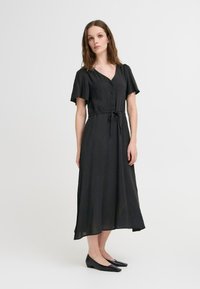Abito maxi nero con maniche a uccello corte, scollatura a V, bottoni frontali, vita cintrata e silhouette fluida, abbinato a scarpe basse nere.