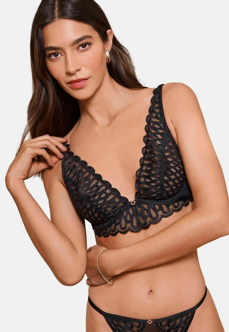 Mujer con cabello largo y oscuro que lleva un bralette de encaje negro y ropa interior a juego, complementado con pendientes de aro dorados y una pulsera.