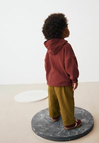 Un sweat à capuche bordeaux avec une capuche et des épaules tombantes, associé à un pantalon en velours côtelé vert olive. Debout sur une plateforme grise circulaire.