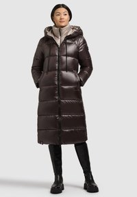 Brauner Steppmantel mit Kapuze, glänzender Textur, vorderem Reißverschluss und elastischen Kordelzügen. Trägt sich über einer hellen Innenjacke.