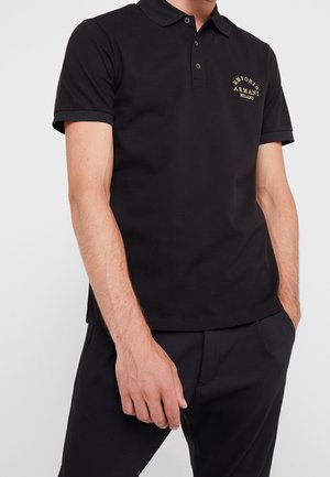 Polo shirt noir avec un col contrasté, des manches courtes et un logo doré brodé sur la poitrine gauche. Texture de tissu lisse et uni.