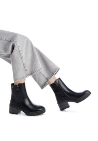 Botas de tobillo de cuero negro con un tacón grueso, cremalleras laterales y suelas texturizadas, combinadas con unos pantalones de mezclilla recortados en gris claro.