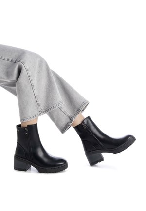 Botas de tobillo de cuero negro con un tacón grueso, cremalleras laterales y suelas texturizadas, combinadas con unos pantalones de mezclilla recortados en gris claro.