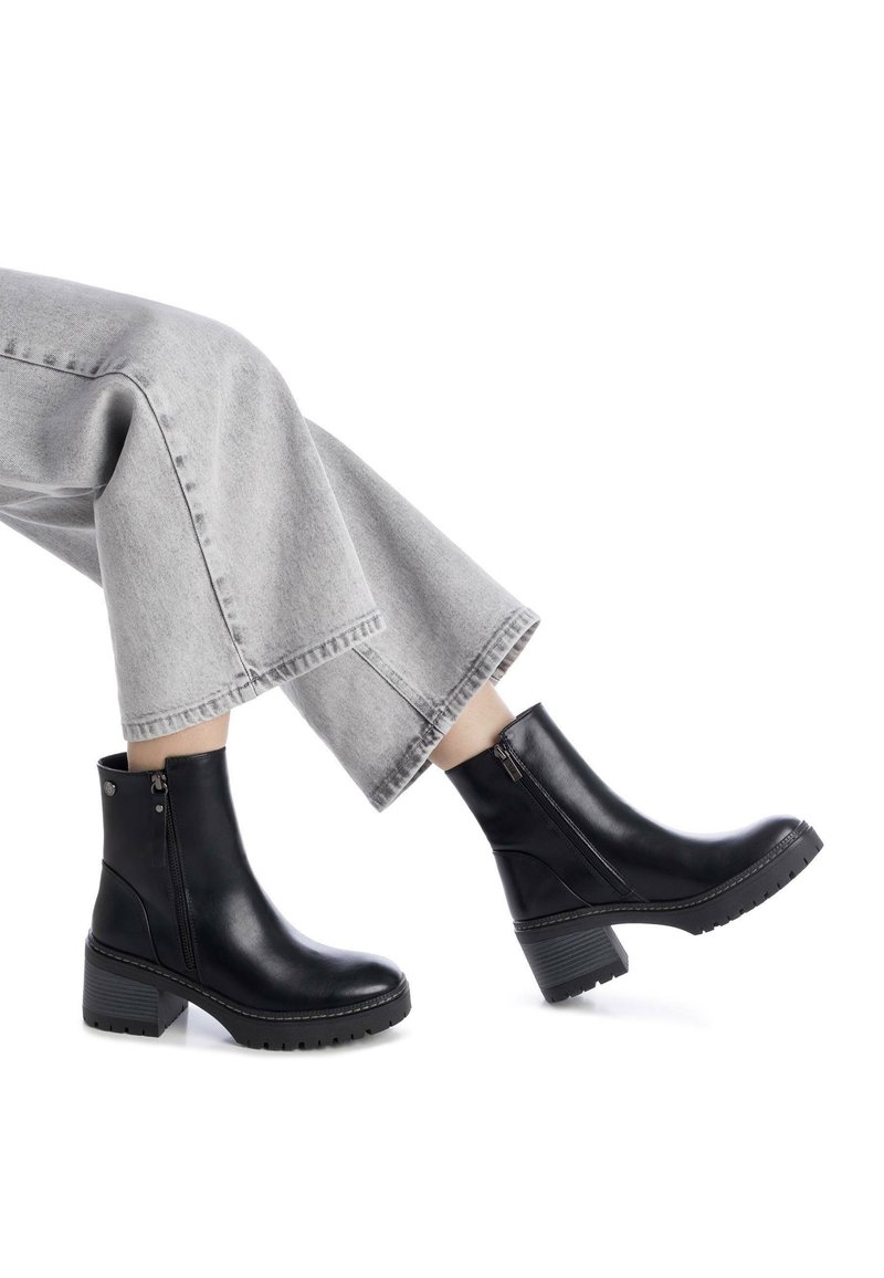 Botas de tobillo de cuero negro con un tacón grueso, cremalleras laterales y suelas texturizadas, combinadas con unos pantalones de mezclilla recortados en gris claro.