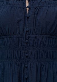 Κοντινή λήψη σε ένδυμα ναυτικού μπλε με κατακόρυφη σειρά κουμπιών καλυμμένων με ύφασμα και ραφές με ελαστικό που δημιουργούν υφασμάτινες, υφασματικές ενότητες.