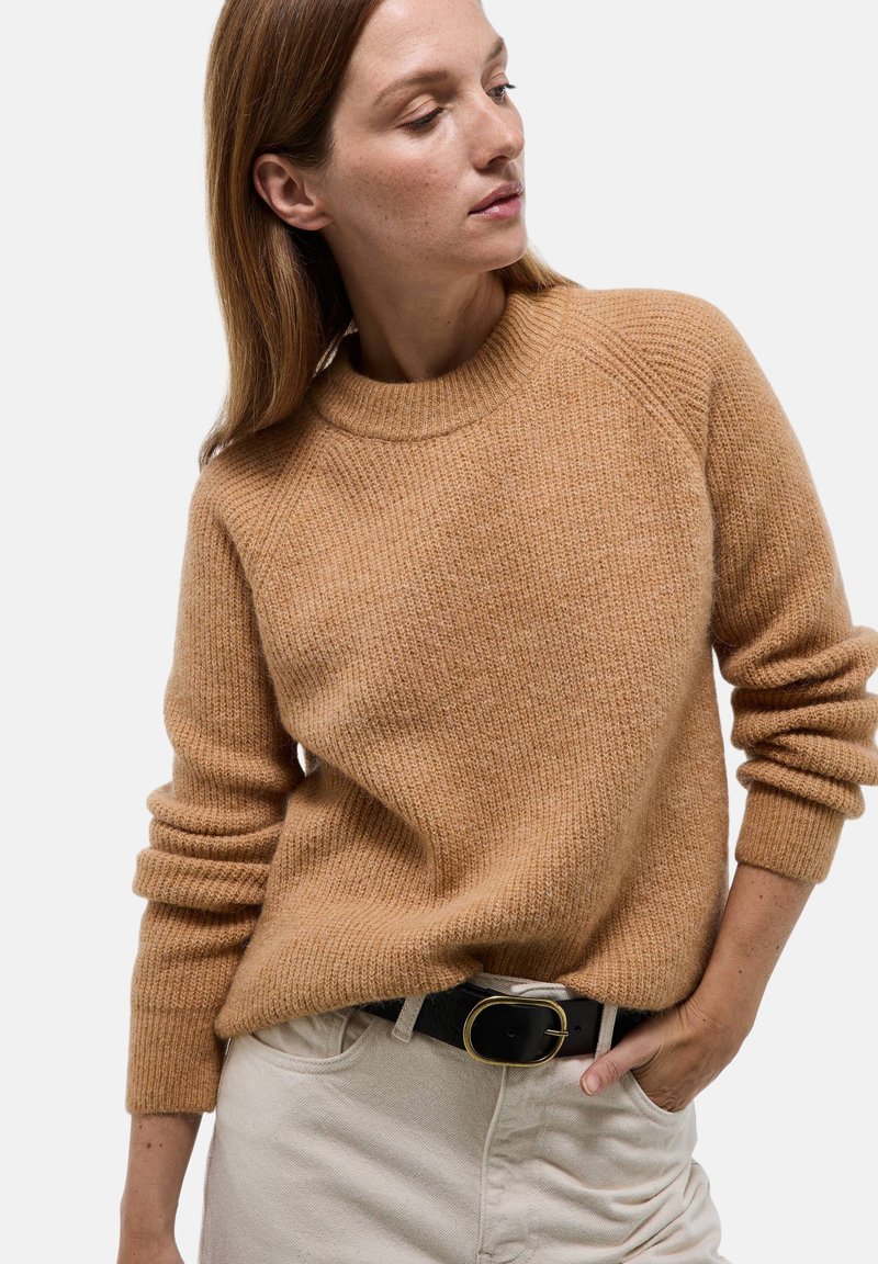 Pull en tricot couleur camel avec un col rond, texture côtelée et poignets roulés, associé à un pantalon clair et une ceinture noire avec une boucle en or.