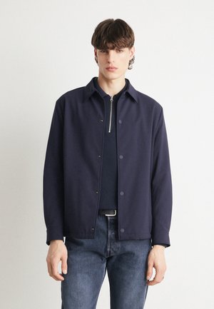Veste bleu marine à boutons avec un col, en tissu doux, et une fermeture éclair sur le devant. Portée sur une chemise sombre et associée à un jean bleu.