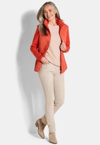 Rode gewatteerde vest met hoge kraag over een beige trui, gecombineerd met lichtbeige pantalons en tan enkellaarsjes. Soepele texturen en een getailleerd ontwerp.